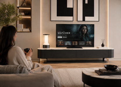 A woman sits on a sofa in a modern living room, watching a large flat-screen TV displaying the Watcher streaming service interface. The TV is mounted above a dark media console in a warm, contemporary interior with wall art, shelving, and ambient lighting.