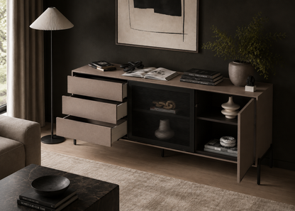A modern sideboard cabinet in a styled living room setting. The cabinet features a light taupe wood finish, three open drawers on the left, a central black wire-grid storage compartment, and open shelves on the right.