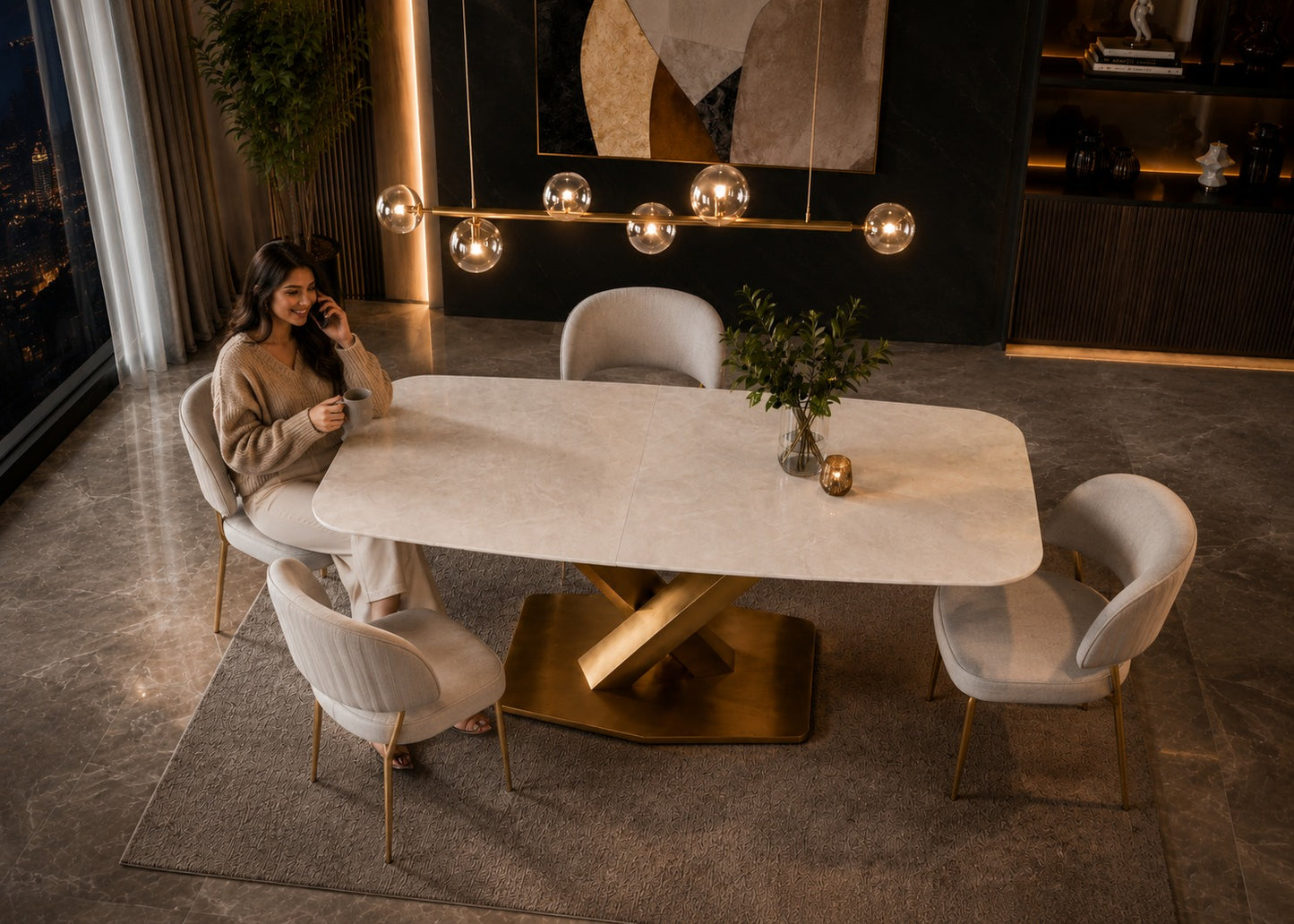 A modern dining room scene with a woman seated at a rectangular white dining table with a gold-tone base, surrounded by four light gray upholstered chairs. The room has warm ambient lighting, a hanging glass globe chandelier, and a dark accent wall.