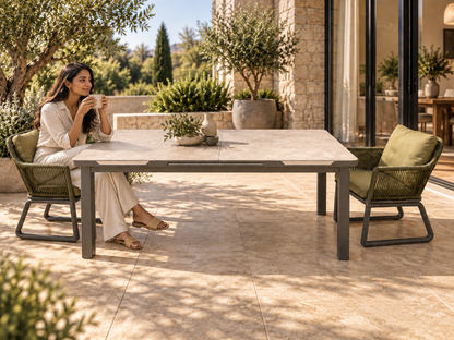 A woman sits at a rectangular outdoor dining table with two woven green chairs on a sunlit patio. The table has a light stone top and dark metal legs, with a stone building, plants, and large windows in the background.
