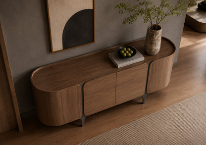 A modern curved wooden sideboard or console cabinet in a warm medium brown wood finish, styled in a minimalist room with a textured gray wall, abstract wall art, a ceramic vase with greenery, and a bowl of green olives on top.
