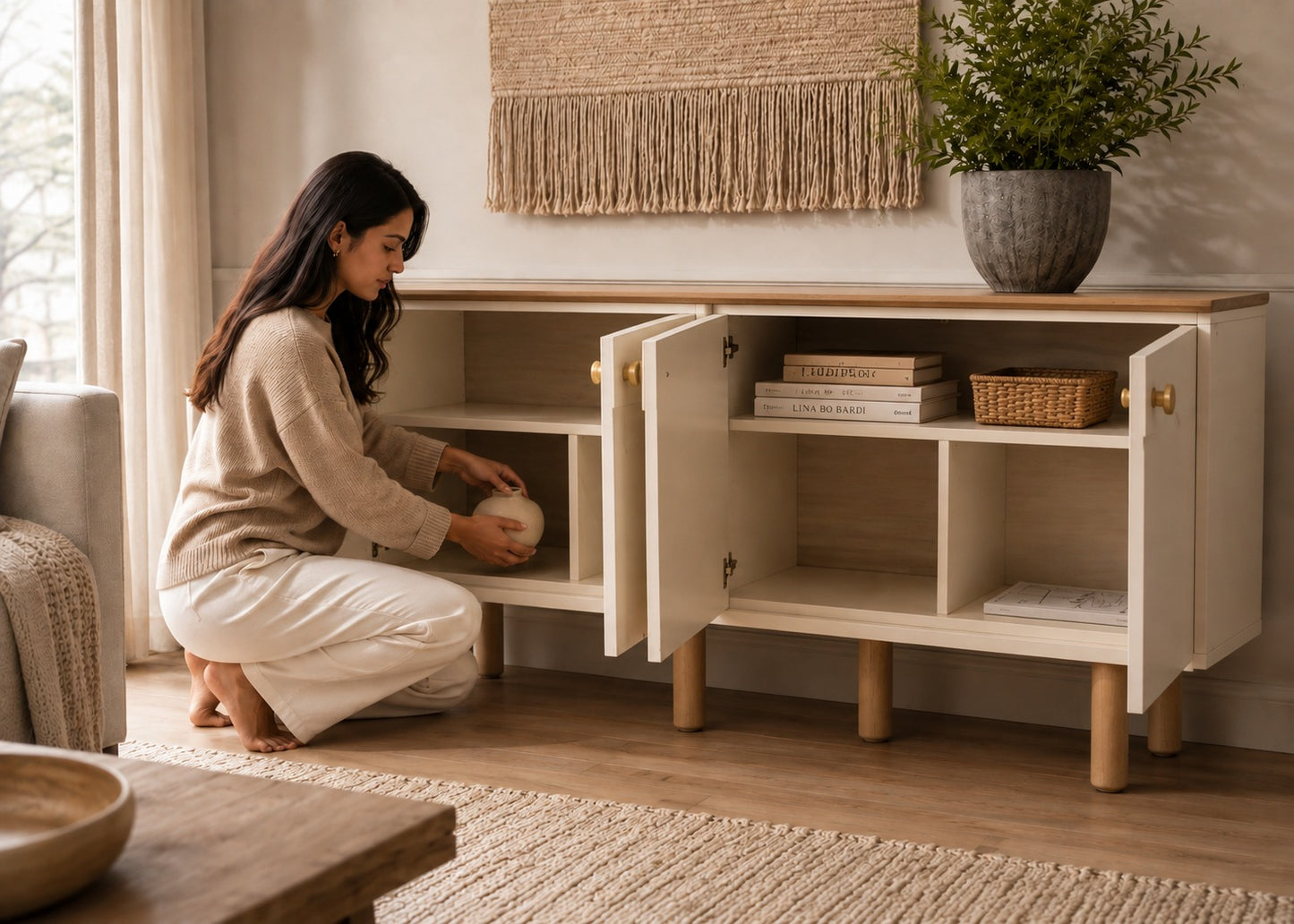 A woman kneels beside a light wood media console in a bright living room, placing a decorative object on one of the open shelves. The cabinet has two doors with small gold knobs, open cubbies, and light wood legs, with books and a basket inside.