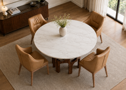 A round white marble dining table with a dark wood pedestal base, surrounded by four tan leather armchairs on a light beige rug in a bright room.