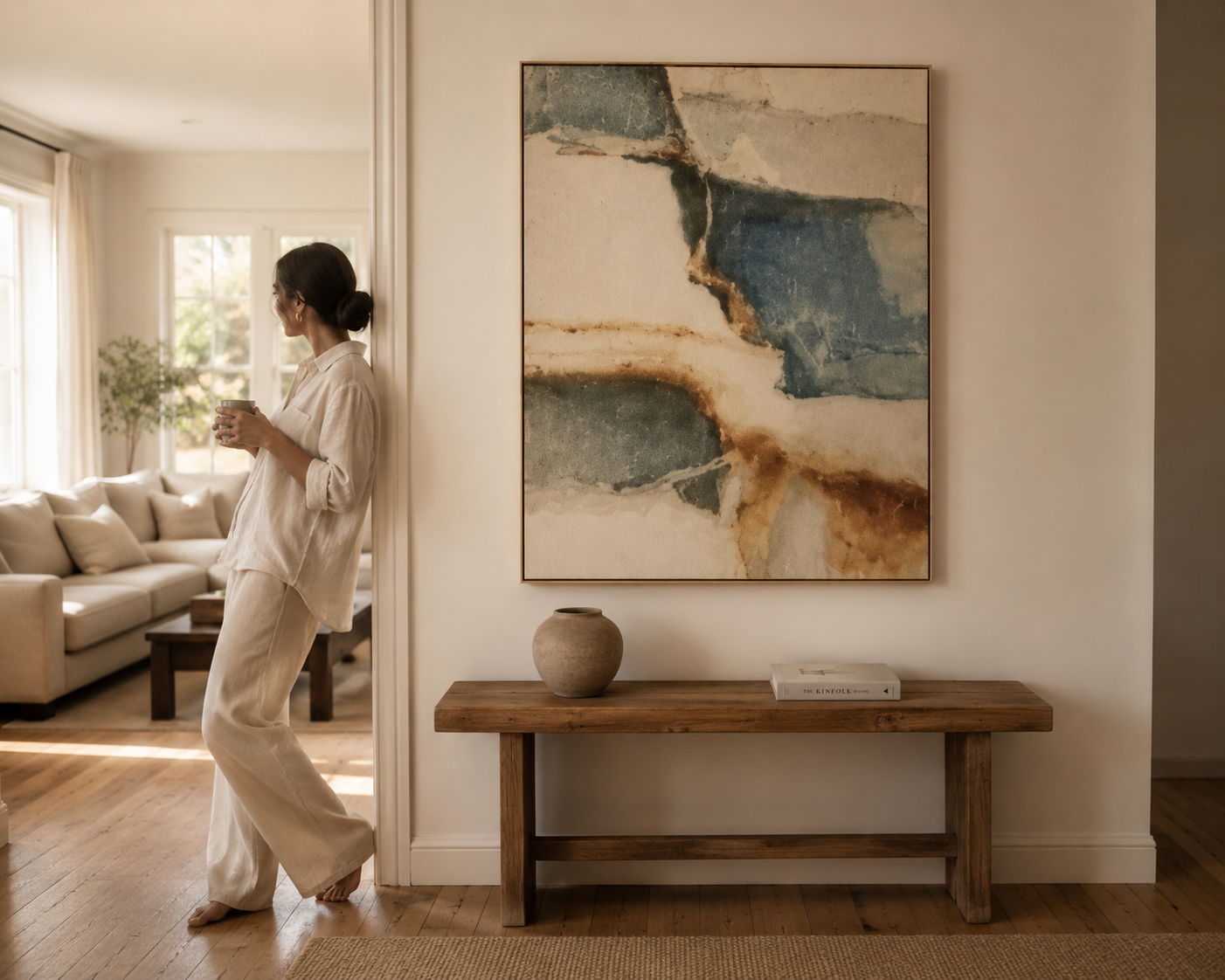 A woman in a relaxed cream-colored outfit leans against a doorway in a bright, modern living room, looking at a large framed abstract painting on the wall. The painting features blue, beige, and brown watercolor-like textures.