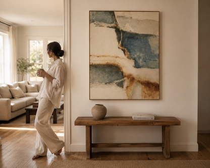 A woman in a relaxed cream-colored outfit leans against a doorway in a bright, modern living room, looking at a large framed abstract painting on the wall. The painting features blue, beige, and brown watercolor-like textures.