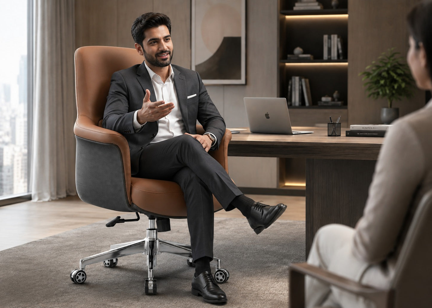 A man in a dark gray suit sits in a modern brown and gray office chair, gesturing while speaking to another person in a professional office setting with a desk, laptop, and shelving in the background.
