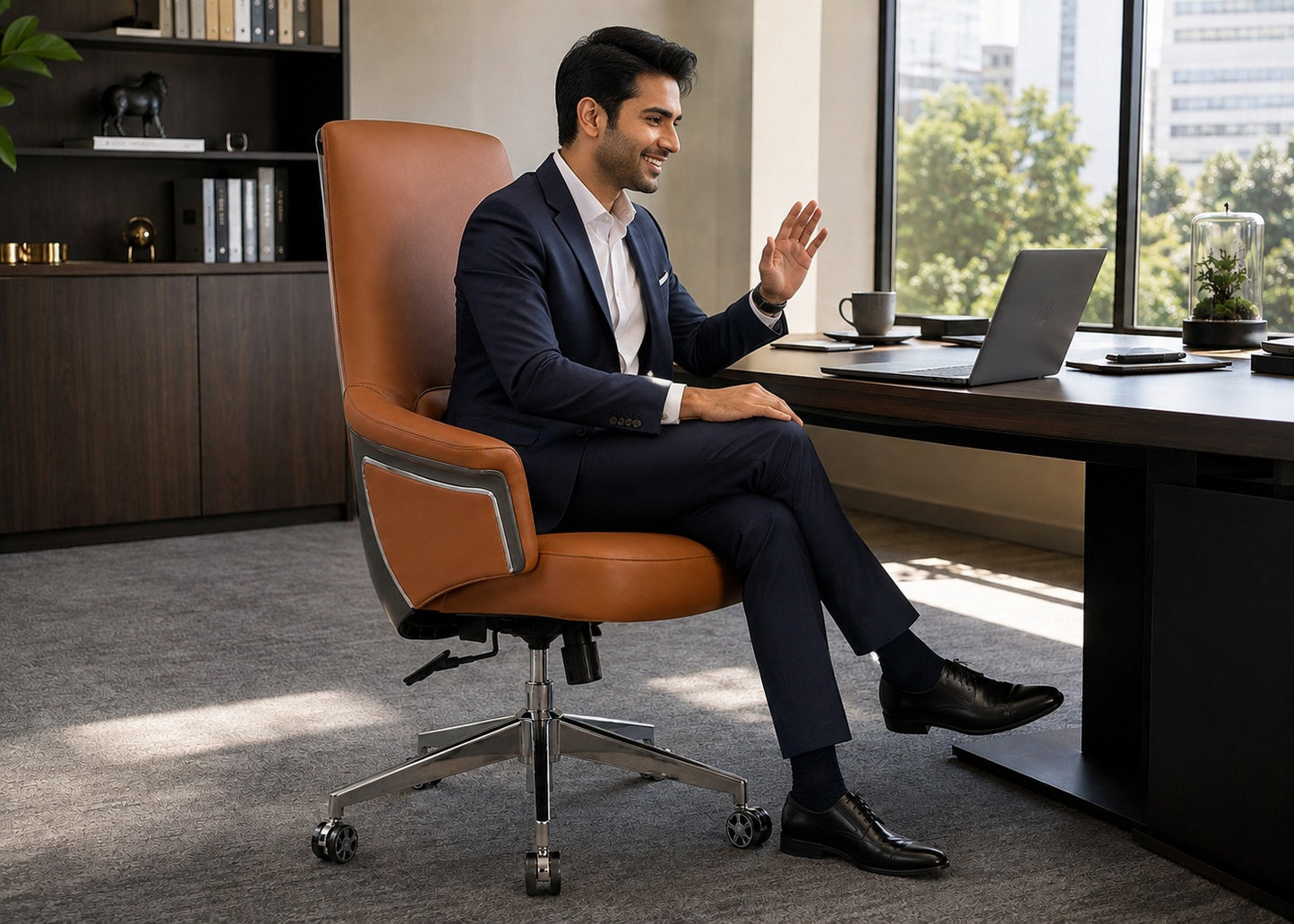 A man in a navy blue suit sits in a brown leather office chair next to a desk with a laptop, in a modern office setting.
