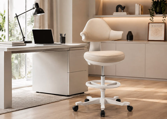 A cream-colored ergonomic office chair with a white wheeled base, positioned next to a matching cream desk in a modern, sunlit home office setting.