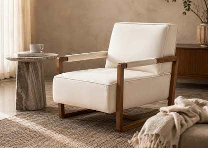 A modern accent chair with cream-colored upholstery and a medium brown wooden frame, styled in a warm, neutral-toned living room setting next to a marble side table.