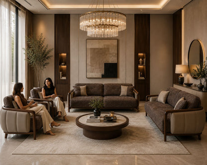 A luxury modern living room featuring a dark brown textured sofa, a matching armchair with two women sitting in it, a second sofa on the right, a round wooden coffee table, and a large tiered crystal chandelier hanging from the ceiling.